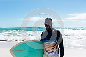 African american retired senior man carrying surfboard at beach against sky on sunny day