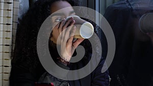 African American sad girl teenager young woman teen using her mobile cell phone looking out of a window and drinking cup of coffee