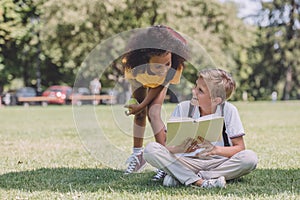 African american schoolboy standing near multicultural friend sitting on lawn with book