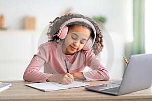 African american schoolgirl having online lesson, using laptop and headphones at home, taking notes