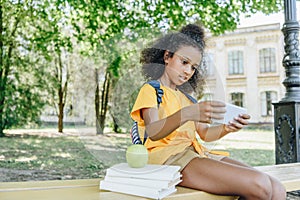 African american schoolgirl sitting on bench near books and using smartphone