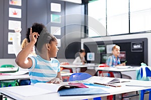 African american schoolgirl sitting at desk raising hand in elementary school classroom, copy space