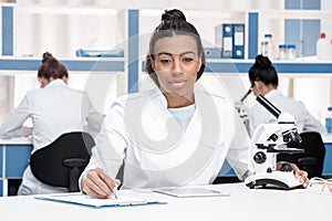 African american scientist in lab coat with clipboard, microscope and digital tablet working in chemical lab