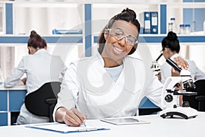 African american scientist in lab coat with clipboard, microscope and digital tablet working in chemical lab