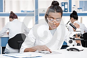 African american scientist in lab coat with microscope and digital tablet working in chemical lab