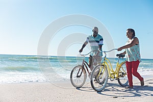 African american senior couple wheeling bicycles at beach against blue sky with copy space