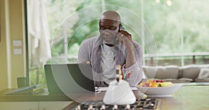 African american senior man in bathrobe sitting in kitchen using laptop and talking on smartphone