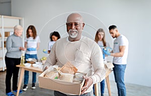 African american senior man holding box with donations food, looking and smiling at camera in charity center