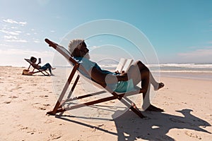 African american senior man and mature woman reading books while relaxing on deckchairs at beach