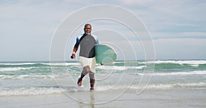 African american senior man walking on a beach holding surfboard and running out of the sea