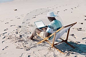 African american senior man wearing hat working over laptop while relaxing on deckchair at beach
