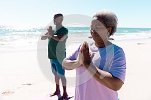 African american senior man and woman meditating in prayer pose at beach against clear sky