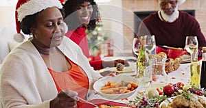 African american senior woman in santa hat serving food in plate while sitting on dining table havin