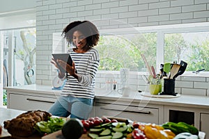 African American smiling female researching recipes on digital tablet leaning against counter in clean kitchen.