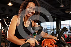 African American smiling woman doing cardio workout on exercise bike in gym smiling at camera.