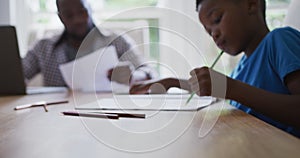 African american son sitting at kitchen table doing school work with father working in background