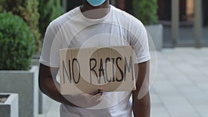 An African American stands in the middle of the street holding a cardboard poster in front of him that reads NO RACISM