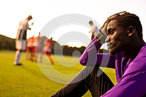 African american stressed male goalkeeper with head in hand sitting on field against clear sky