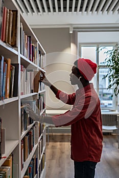 African American student man picking up book from shelf while standing between library bookcases