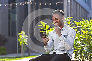 African american student man successful reads the results of his work online from the phone, outdoors, smiles and rejoices