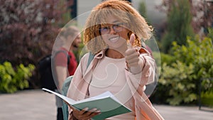 African American student woman reading book outdoors smiling at camera showing thumb up high school pupil schoolgirl