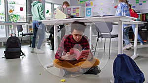 African-American teen student read book sitting under desk in noisy class