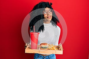 African american woman with afro hair eating a tasty classic burger with fries and soda smiling looking to the side and staring