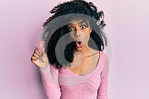 African american woman with afro hair holding menstrual cup scared and amazed with open mouth for surprise, disbelief face