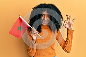 African american woman with afro hair holding morocco flag doing ok sign with fingers, smiling friendly gesturing excellent symbol