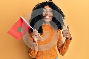 African american woman with afro hair holding morocco flag smiling happy and positive, thumb up doing excellent and approval sign