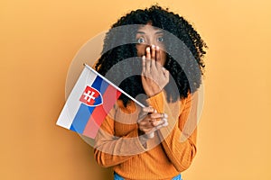 Afroameričanka s afro vlasmi drží slovenskú vlajku zakrývajúcu ústa rukou, šokovaná a bojí sa chyby