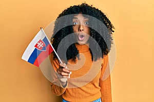 Afroameričanka s afro vlasy držící slovenskou vlajku vyděšená a ohromená s otevřenými ústy k překvapení, nevěřícná tvář