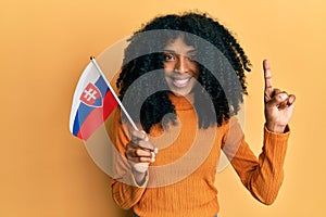 Afroameričanka s afro vlasmi drží vlajku slovenska s úsmevom s myšlienkou alebo otázkou ukazovákom so šťastnou tvárou,