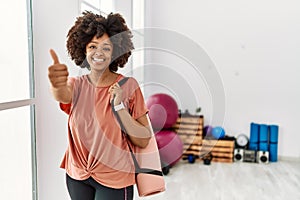 African american woman with afro hair holding yoga mat at pilates room approving doing positive gesture with hand, thumbs up
