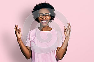 African american woman with afro hair wearing casual clothes and glasses gesturing finger crossed smiling with hope and eyes
