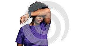 African american woman with afro hair wearing casual purple t shirt covering eyes with arm, looking serious and sad