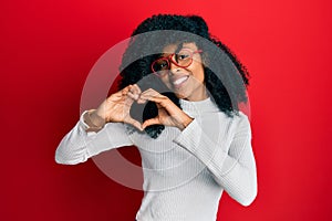 African american woman with afro hair wearing casual sweater and glasses smiling in love doing heart symbol shape with hands