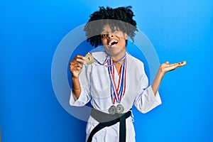 African american woman with afro hair wearing karate kimono and black belt holding medals celebrating achievement with happy smile