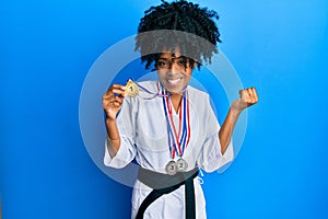 African american woman with afro hair wearing karate kimono and black belt holding medals screaming proud, celebrating victory and