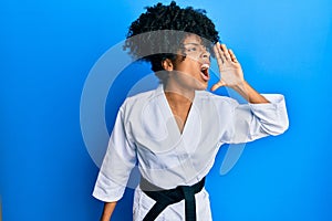 African american woman with afro hair wearing karate kimono and black belt shouting and screaming loud to side with hand on mouth