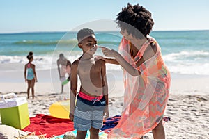 African american woman applying lotion on son's face while father and daughter playing at beach