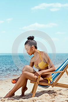 african american woman applying sunscreen lotion on skin while sitting on deck chair on sandy