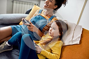 African american woman, baby sitter and caucasian cute little girl having fun together, playing video games, sitting on