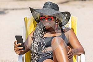 African-American woman on the beach using phone relaxing in deck chair wearing a black hat and sunglasses