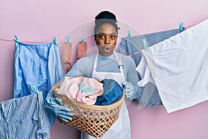 African american woman with braided hair doing laundry holding wicker basket scared and amazed with open mouth for surprise,