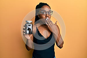 African american woman with braided hair holding jar with coffee beans covering mouth with hand, shocked and afraid for mistake