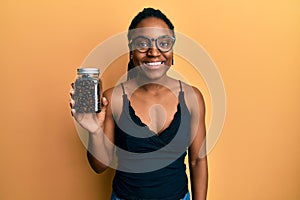 African american woman with braided hair holding jar with coffee beans looking positive and happy standing and smiling with a