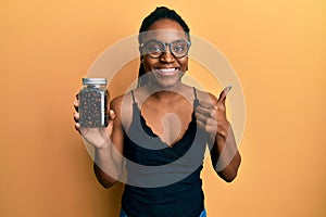 African american woman with braided hair holding jar with coffee beans smiling happy and positive, thumb up doing excellent and