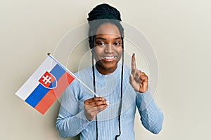 Afroameričanka so zapletenými vlasmi drží slovenskú vlajku s úsmevom s myšlienkou alebo otázkou ukazovákom so šťastnou tvárou,