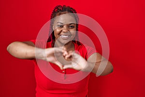 African american woman with braided hair standing over red background smiling in love doing heart symbol shape with hands